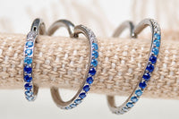 Three silver-toned hoop earrings with rows of blue and light blue rhinestones, displayed on a beige woven ring stand. Pink text at the bottom reads 'Gold, Rose Gold, Purple, Silver Choose your custom metal color'.