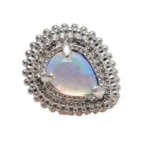 A teardrop-shaped silver ring featuring a central iridescent opal-like stone surrounded by a halo of small clear stones and a textured beaded outer border, displayed on a pale stone surface against a white background.