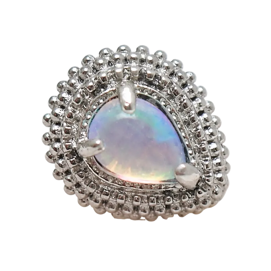 A teardrop-shaped silver ring featuring a central iridescent opal-like stone surrounded by a halo of small clear stones and a textured beaded outer border, displayed on a pale stone surface against a white background.