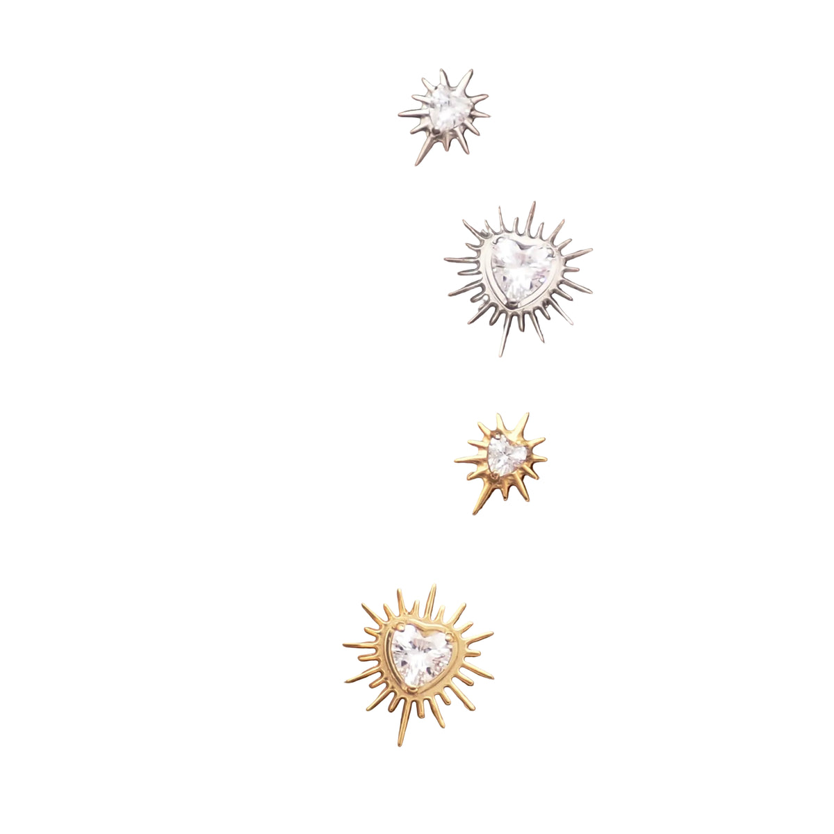 Four small stud earrings displayed on a brown textured card. Two earrings are gold-toned sunburst shapes with clear heart-shaped stones, and two are silver-toned sunburst shapes with clear heart-shaped stones.