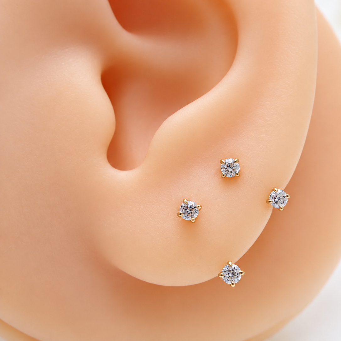 Four small gold stud earrings with clear gemstones displayed on a peach-colored ear mannequin against a soft white background.